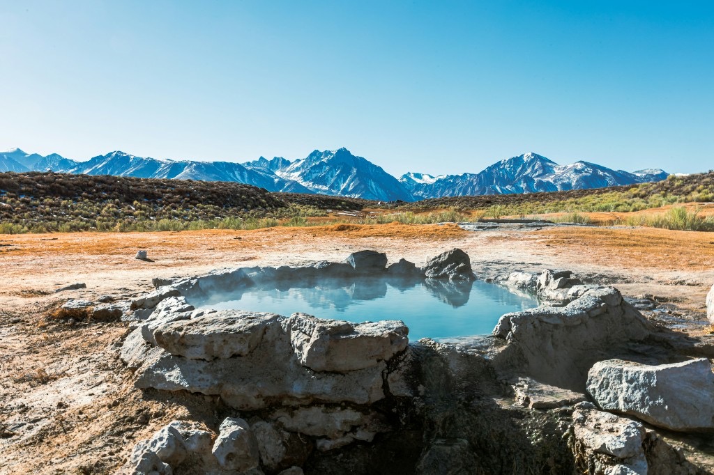 A natural hot spring with steam rising, backed by open scrubland and snow-capped mountains